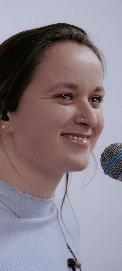 Das Bild zeigt eine junge Frau mit braunem Haar und einem Nasenpiercing, die lächelnd vor einem hellen Hintergrund zu sehen ist. Sie trägt ein weißes Oberteil und steht vor einem Mikrofon, was darauf hindeutet, dass sie spricht oder singt.