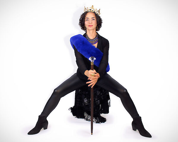 Eine Frau sitzt selbstbewusst vor weißem Hintergrund und trägt eine goldene Krone, einen leuchtend blauen Pelzschal sowie ein farbenfröhliches Perlencollier über einem schwarzen Outfit. In den Händen hält sie einen eleganten Gehstock mit einem Kristallknauf und posiert in einer ausdrucksstarken, königlichen Haltung.