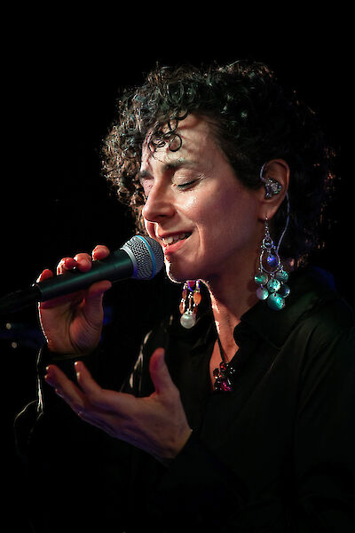 Eine Frau singt in ein Mikrofon.
Sie hat lockiges Haar.
Ihre Augen sind geschlossen.
Sie singt mit viel Gefühl.
Mit einer Hand macht sie eine Geste.
Sie trägt türkisfarbene Ohrringe.
Sie trägt einen kleinen Ohrhörer.
Hinter ihr ist es dunkel.
Ein Licht beleuchtet sie.