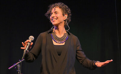 Eine Frau steht auf einer Bühne. Sie lächelt. Sie hält ein Mikrofon in der Hand. Sie streckt den anderen Arm aus. Sie trägt schwarze Kleidung. Sie trägt ein blaues Perlenhalsband. Sie hat einen kleinen Hörer im Ohr. Die Frau tritt live auf.