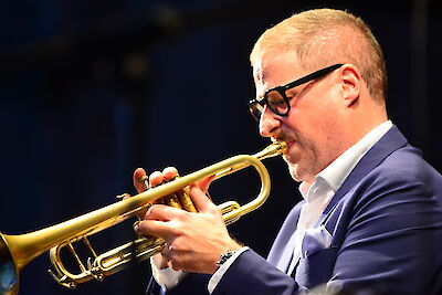 A man with black glasses and a blue jacket is concentrating on playing a golden trumpet. The musician is seen in profile as he holds his instrument in front of a dark blue background during a performance.