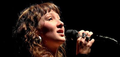 Eine junge Frau singt auf einer Bühne. Sie hat lockiges braunes Haar. Sie trägt Kreolen-Ohrringe. Sie hält ein Mikrofon in der Hand. Ihre Augen sind geschlossen. Das Licht auf der Bühne macht die Stimmung besonders.