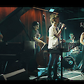 Eine Jazzband tritt auf einer Bühne auf, bestehend aus einer Sängerin am Mikrofon, einem Pianisten, einem Kontrabassisten, einem Schlagzeuger und einer Saxophonistin. Die Musizierenden sind in einem abgedunkelten Veranstaltungsraum zu sehen, der von farbigem Bühnenlicht beleuchtet wird.
