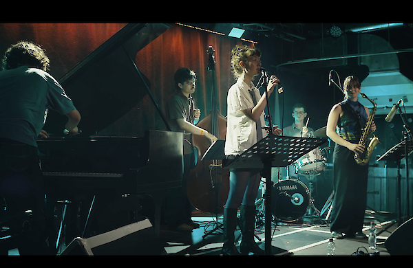 Eine Jazzband tritt auf einer Bühne auf, bestehend aus einer Sängerin am Mikrofon, einem Pianisten, einem Kontrabassisten, einem Schlagzeuger und einer Saxophonistin. Die Musizierenden sind in einem abgedunkelten Veranstaltungsraum zu sehen, der von farbigem Bühnenlicht beleuchtet wird.