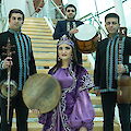 Eine Gruppe von vier Musikerinnen und Musikern in traditionellen aserbaidschanischen oder zentralasiatischen Kostümen posiert auf einer modernen Treppe und hält traditionelle Instrumente wie Rahmentrommeln (Def/Qaval), eine Kemancha (Streichinstrument) und eine Saz (Langhalslaute) in den Händen. Die Frau in der Mitte trägt ein auffälliges lila Gewand mit einer Krone und hält ebenfalls eine große Rahmentrommel, während die Männer in schwarz-türkisfarbenen Kostümen gekleidet sind.