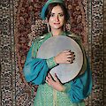 Eine junge Frau in einem eleganten türkisfarbenen traditionellen Kostüm mit goldenen Verzierungen hält eine Rahmentrommel (Qaval/Def) in den Armen und lächelt in die Kamera. Im Hintergrund ist ein reich gemusterter orientalischer Teppich vor einer Backsteinwand zu sehen, was auf einen kulturellen oder folkloristischen Kontext aus dem aserbaidschanischen oder zentralasiatischen Raum hindeutet.