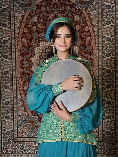 Eine junge Frau in einem eleganten türkisfarbenen traditionellen Kostüm mit goldenen Verzierungen hält eine Rahmentrommel (Qaval/Def) in den Armen und lächelt in die Kamera. Im Hintergrund ist ein reich gemusterter orientalischer Teppich vor einer Backsteinwand zu sehen, was auf einen kulturellen oder folkloristischen Kontext aus dem aserbaidschanischen oder zentralasiatischen Raum hindeutet.