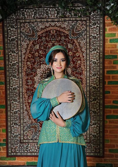 Eine junge Frau in einem eleganten türkisfarbenen traditionellen Kostüm mit goldenen Verzierungen hält eine Rahmentrommel (Qaval/Def) in den Armen und lächelt in die Kamera. Im Hintergrund ist ein reich gemusterter orientalischer Teppich vor einer Backsteinwand zu sehen, was auf einen kulturellen oder folkloristischen Kontext aus dem aserbaidschanischen oder zentralasiatischen Raum hindeutet.