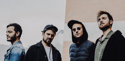 Vier junge Männer stehen nebeneinander vor einem urbanen Hintergrund aus Beton und Ziegelsteinen. Sie tragen lässige Kleidung, darunter Caps und eine Sonnenbrille, und blicken in verschiedene Richtungen – das Bild wirkt wie ein professionelles Bandfoto.