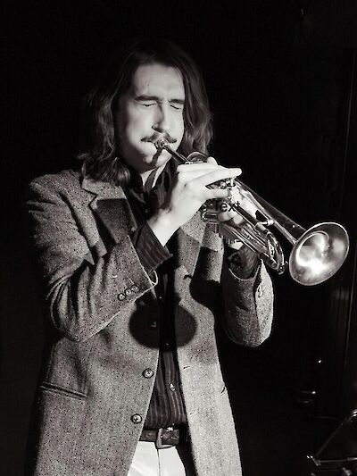 Ein junger Mann mit langen Haaren und einem Tweed-Jacket spielt leidenschaftlich Trompete, die Augen geschlossen. Das Schwarz-Weiß-Foto vermittelt eine stimmungsvolle, jazzige Atmosphäre einer Live-Performance.