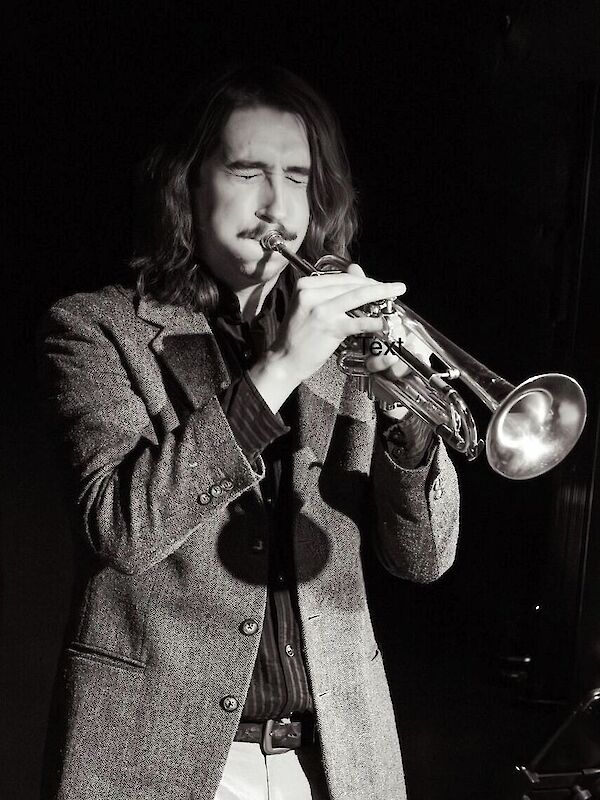 Ein junger Mann mit langen Haaren und einem Tweed-Jacket spielt leidenschaftlich Trompete, die Augen geschlossen. Das Schwarz-Weiß-Foto vermittelt eine stimmungsvolle, jazzige Atmosphäre einer Live-Performance.