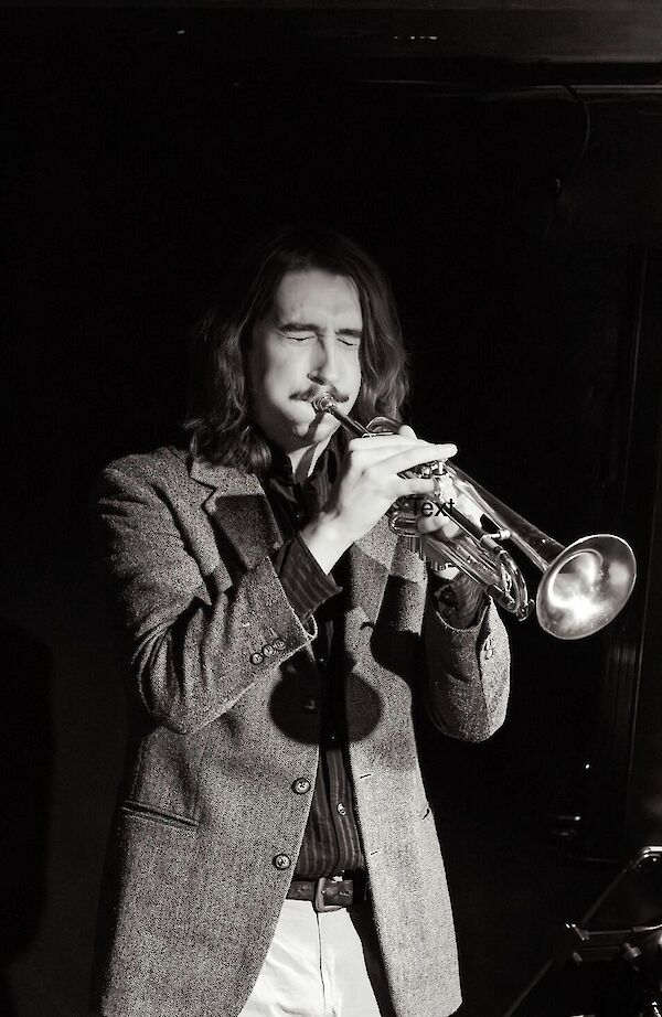A young man with long hair and a tweed jacket plays the trumpet with concentration, his eyes closed. The black-and-white photo conveys the moody, jazzy atmosphere of a live performance.