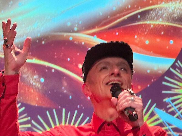 Eine Person in einem roten Outfit singt leidenschaftlich in ein Mikrofon und hebt dabei eine Hand gen Himmel. Im Hintergrund ist eine farbenprächtige, psychedelische Projektion mit leuchtenden Farben und Mustern zu sehen.