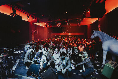 Eine große Gruppe von Musikerinnen und Musikern posiert auf einer Konzertbühne für ein gemeinsames Foto, während im Hintergrund eine begeisterte Menge von Zuschauerinnen und Zuschauern zu sehen ist. Die Veranstaltungshalle ist in warmes rotes Licht getaucht, und rechts im Bild ist eine weiße Pferdeskulptur zu erkennen.