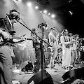 Eine große Band performs live auf einer Bühne, wobei mehrere Musikerinnen und Musiker mit verschiedenen Instrumenten wie Gitarre, Tuba, Trompete und Saxofon zu sehen sind. Das schwarz-weiße Foto zeigt die Gruppe im „domicil" in Dortmund, beleuchtet von Scheinwerfern, die eine dynamische Konzertatmosphäre erzeugen.