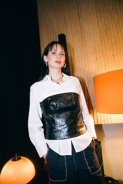 Eine junge Frau steht vor einer Holzwand. Sie hat dunkle Haare. Sie trägt ein weißes Hemd. Darüber trägt sie ein schwarzes Korsett aus Leder. Sie trägt dunkle Hosen. Sie trägt Schmuck aus Silber. Neben ihr stehen zwei Lampen. Die Lampen leuchten warm.