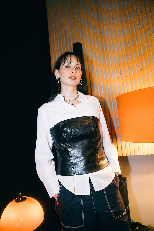 Eine junge Frau mit dunklen Haaren steht vor einer strukturierten Holzwand und trägt ein stilvolles Outfit bestehend aus einem weißen Hemd, einem schwarzen Lederkorsett und dunklen Hosen. Sie ist mit Silberschmuck accessoirisiert, und neben ihr sind zwei Lampen mit warmem Licht zu sehen.