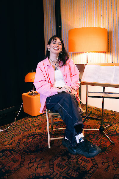 Eine junge Frau sitzt auf einem Stuhl. Sie lächelt. Sie hat dunkle Haare. Sie trägt ein rosa Hemd. Das Hemd ist groß. Sie trägt dunkle Jeans. Sie trägt schwarze Schuhe. Die Schuhe haben eine dicke Sohle. Sie sitzt in einem Musik-Studio. Im Hintergrund steht ein Notenständer. Darauf liegen Noten. Es gibt einen orangen Teppich. Es gibt orange Lampen.