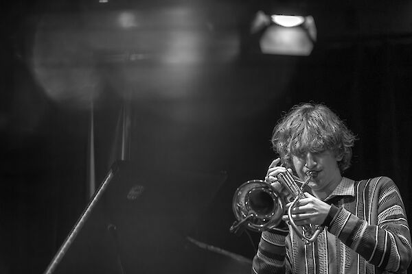 A young musician with curly hair concentrates on playing the trumpet on a stage. The black and white photo shows him in the spotlight, with stage equipment and darkness visible in the background.
