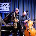 Four musicians pose on the stage of the Jazzschmiede Düsseldorf, two of them holding their instruments - a saxophone and a double bass - in their hands. A grand piano and the Jazzschmiede Düsseldorf logo can be seen in the background.