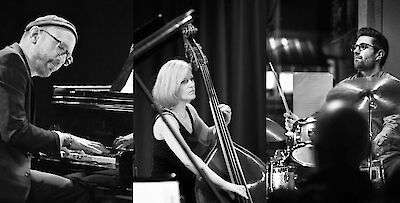 Ein Jazz-Trio in Schwarz-Weiß-Fotografie: Ein älterer Mann spielt Klavier, eine Frau spielt Kontrabass, und ein junger Mann mit Brille sitzt am Schlagzeug. Die drei Musiker sind in konzentrierter Spielhaltung zu sehen und erwecken den Eindruck eines professionellen Konzertauftritts.