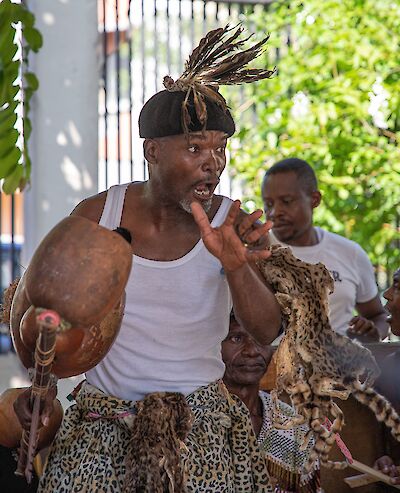 Ein Mann in traditioneller afrikanischer Kleidung mit einem Federschmuck auf dem Kopf und einem Leopardenfell um die Hüften führt leidenschaftlich eine rituelle Zeremonie oder einen Tanz auf, während er ein Kalebassen-Instrument hält. Im Hintergrund sind weitere Personen zu sehen, die an der Veranstaltung teilnehmen.
