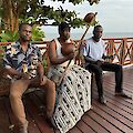 Drei Männer sitzen auf einer Holzterrasse am Meer und musizieren mit traditionellen afrikanischen Instrumenten, darunter eine Kora oder ähnliche Saiteninstrumente aus Kalebassen. Im Hintergrund sind Bäume und der Ozean unter einem bewölkten Himmel zu sehen.