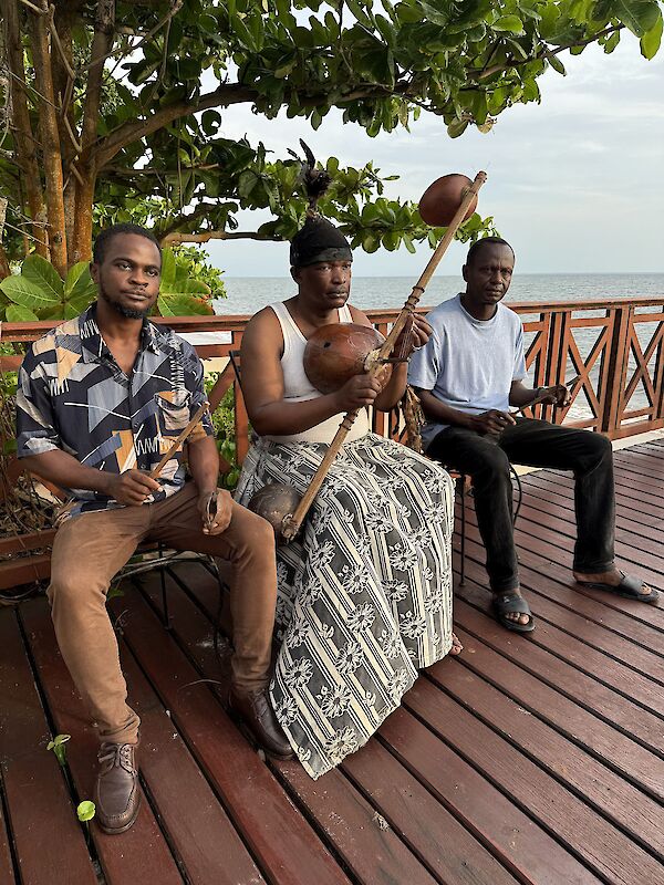 Drei Männer sitzen auf einer Holzterrasse am Meer und musizieren mit traditionellen afrikanischen Instrumenten, darunter eine Kora oder ähnliche Saiteninstrumente aus Kalebassen. Im Hintergrund sind Bäume und der Ozean unter einem bewölkten Himmel zu sehen.
