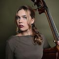 Eine junge Frau mit langen, leicht gewellten Haaren lehnt nachdenklich an einem Kontrabass und hält das Instrument mit einer Hand fest. Das professionelle Porträtfoto zeigt sie vor einem olivgrünen Hintergrund in einem schlichten grauen Oberteil.