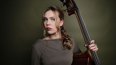 Eine junge Frau mit langen, leicht gewellten Haaren lehnt nachdenklich an einem Kontrabass und hält das Instrument mit einer Hand fest. Das professionelle Porträtfoto zeigt sie vor einem olivgrünen Hintergrund in einem schlichten grauen Oberteil.