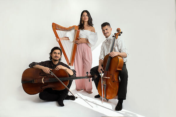 Drei junge Musikerinnen und Musiker posieren vor einem weißen Hintergrund: zwei Cellisten und eine Harfenistin in der Mitte. Die Gruppe präsentiert sich in einem professionellen Fotoshooting, gekleidet in elegante, helle Outfits.