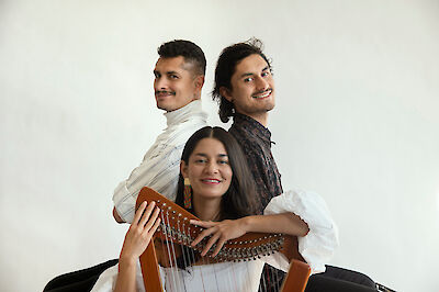 Drei junge Musikerinnen und Musiker posieren zusammen vor einem weißen Hintergrund, wobei eine Frau im Vordergrund eine kleine Harfe hält. Die zwei Männer stehen hinter ihr Rücken an Rücken und alle drei lächeln in die Kamera.