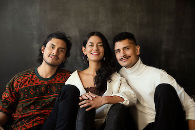 Drei junge Menschen – zwei Männer und eine Frau in der Mitte – sitzen eng beieinander vor einem dunklen Hintergrund und lächeln freundlich in die Kamera. Sie tragen lässige, stilvolle Kleidung und wirken entspannt bei diesem professionellen Porträtfoto.