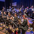 Eine Big Band tritt auf einer Bühne der „Jazzschmiede" in Düsseldorf auf, wobei die Musikerinnen und Musiker überwiegend in Schwarz gekleidet sind. Im Vordergrund spielen mehrere Saxofonistinnen, während im Hintergrund Posaunisten und Trompeter zu sehen sind.
