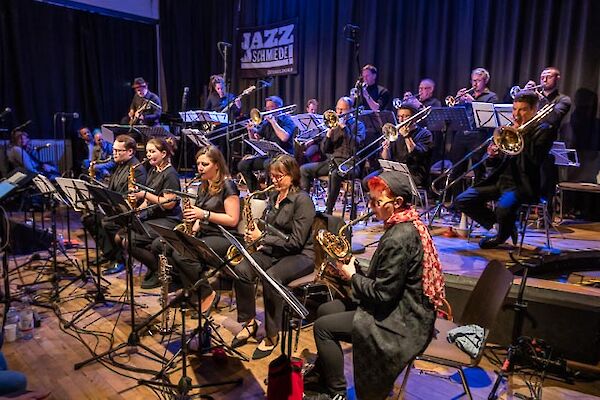 Eine Big Band tritt auf einer Bühne der „Jazzschmiede" in Düsseldorf auf, wobei die Musikerinnen und Musiker überwiegend in Schwarz gekleidet sind. Im Vordergrund spielen mehrere Saxofonistinnen, während im Hintergrund Posaunisten und Trompeter zu sehen sind.