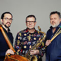Drei Musiker posieren vor einem grauen Hintergrund und halten ihre jeweiligen Instrumente in den Händen: ein Kontrabass, ein Saxofon und Schlagzeugstöcke. Es handelt sich um ein professionelles Portraitfoto, vermutlich einer Jazz-Band oder eines Musikensembles.
