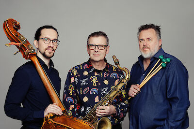 Drei Musiker stehen vor einem grauen Hintergrund. Sie halten ihre Instrumente. Ein Musiker hält einen Kontrabass. Ein anderer hält ein Saxofon. Der dritte hält Schlagzeugstöcke. Das Foto zeigt eine Jazz-Band.