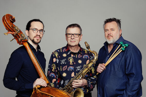 Drei Musiker posieren vor einem grauen Hintergrund und halten ihre jeweiligen Instrumente in den Händen: ein Kontrabass, ein Saxofon und Schlagzeugstöcke. Es handelt sich um ein professionelles Portraitfoto, vermutlich einer Jazz-Band oder eines Musikensembles.