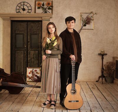 Zwei junge Menschen stehen in einem vintage-dekorierten Raum mit Holzdielen und rustikalen Wanddekorationen – ein junges Mädchen mit langen blonden Haaren hält weiße Lilien in der Hand, während ein junger Mann neben ihr eine klassische Gitarre hält. Im Hintergrund sind eine alte Holztür, eine Wanduhr, gerahmte Blumenbilder sowie ein Kontrabass zu sehen.