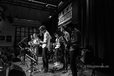Eine Gruppe junger Musikerinnen und Musiker tritt auf einer Bühne der „Jazzschmiede" auf und spielt verschiedene Instrumente, darunter Saxofon und Gitarre. Das Schwarz-Weiß-Foto zeigt das Ensemble vor einem sitzenden Publikum in einem abgedunkelten Veranstaltungsraum.