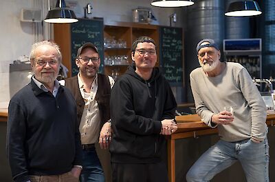 Vier Männer stehen nebeneinander und lächeln in die Kamera, angelehnt an einen Holztresen in einem modernen Bar- oder Weinlokal. Im Hintergrund sind Tafelkreideschriften, Glasregale und Industrielampen zu sehen, die dem Raum eine gemütliche Atmosphäre verleihen.