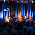 Ein Jazz-Trio mit Klavier, Kontrabass und Schlagzeug tritt auf der Bühne der „Jazzschmiede" auf, während die Musiker durch eine Langzeitbelichtung leicht verschwommen wirken. Ein zahlreiches Publikum verfolgt das Konzert in dem stimmungsvoll blau beleuchteten Veranstaltungsraum.