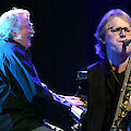 Two middle-aged musicians with grey hair are performing together on stage, one of them playing the piano and the other the tenor saxophone. Both are dressed in dark clothes and are absorbed in their playing, whilst blue stage lighting casts an atmospheric glow over the scene.