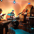 A five-piece band is playing in an atmospheric, dimly lit vaulted cellar bathed in warm orange tones. The line-up consists of a double bassist, a drummer, a guitarist, a vibraphone player and a musician playing a steel guitar or lap steel.