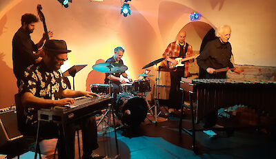 Eine fünfköpfige Band spielt in einem stimmungsvoll beleuchteten Gewölbekeller mit warmen Orangetönen. Zu sehen sind ein Kontrabassist, ein Schlagzeuger, ein Gitarrist, ein Vibraphonspieler sowie ein Musiker an einem Steelgitarre- oder Lap-Steel-Instrument.