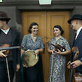 Vier Musikerinnen und Musiker in vintageartiger Kleidung stehen vor einer gelben Holztür und halten traditionelle Instrumente wie Geigen, eine Rahmentrommel und ein Cello in den Händen. Die Gruppe wirkt fröhlich und entspannt, zwei der Personen tragen Hüte, was der Szene einen nostalgischen Charakter verleiht.