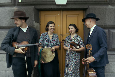 Vier Musikerinnen und Musiker in vintageartiger Kleidung stehen vor einer gelben Holztür und halten traditionelle Instrumente wie Geigen, eine Rahmentrommel und ein Cello in den Händen. Die Gruppe wirkt fröhlich und entspannt, zwei der Personen tragen Hüte, was der Szene einen nostalgischen Charakter verleiht.