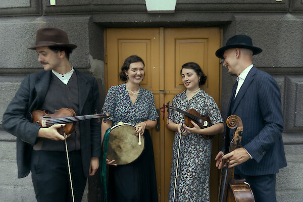 Vier Musikerinnen und Musiker in vintageartiger Kleidung stehen vor einer gelben Holztür und halten traditionelle Instrumente wie Geigen, eine Rahmentrommel und ein Cello in den Händen. Die Gruppe wirkt fröhlich und entspannt, zwei der Personen tragen Hüte, was der Szene einen nostalgischen Charakter verleiht.