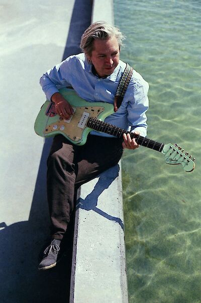 Ein Mann mit hellblondem Haar sitzt auf einer Betonkante am Wasser und spielt eine mintgrüne Fender-Gitarre. Er trägt ein hellblaues Hemd und dunkle Hose, und das türkisfarbene Wasser im Hintergrund verleiht dem Bild eine entspannte, sommerliche Atmosphäre.
