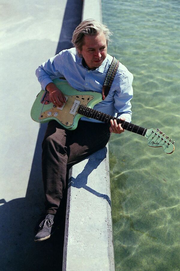 Ein Mann mit hellblondem Haar sitzt auf einer Betonkante am Wasser und spielt eine mintgrüne Fender-Gitarre. Er trägt ein hellblaues Hemd und dunkle Hose, und das türkisfarbene Wasser im Hintergrund verleiht dem Bild eine entspannte, sommerliche Atmosphäre.